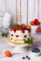 Layered Pavlova dessert on a white pedestal, garnished with mixed berries and mint, presented on a rustic wooden table.