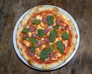 Pizza con salsa roja, queso mozzarella, piña, tocineta y rúgula en un plato blanco sobre un fondo de madera
