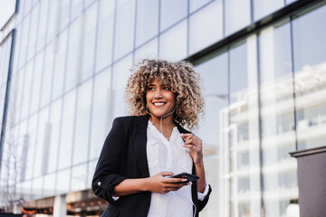 beautiful smiling business woman using mobile phone and headphones in city. Buildings background