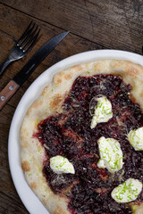 Pizza lista para comer con frutos rojos, queso mozzarella, queso crema, queso parmesano y ralladura de limón en un plato blanco sobre un fondo de madera junto a un cuchillo y un tenedor 