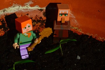 Fototapeta premium LEGO Minecraft figure of Alex with golden shovel and villager in his typical posture checking young nurseling of bamboo from Bambusa genus in large clay pot, spring morning sunshine.