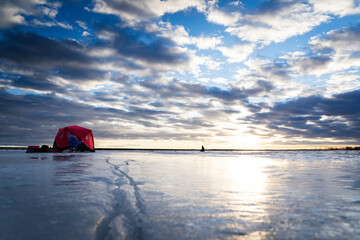 An ice fishing tent on a frozen cracking lake with people fishing under a dramatic sunset in Alberta Canada