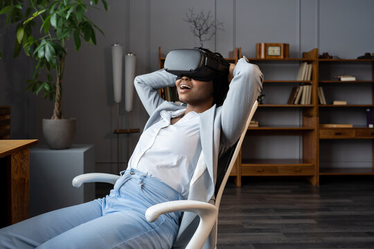 VR for workplace wellness. Amazed african american female employee in virtual reality goggles resting at in relaxing virtual environment, sitting on office chair with hands behind head. AR in business