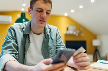 Fototapeta premium Redhead man using smartphone and a credit card in the kitchen at home