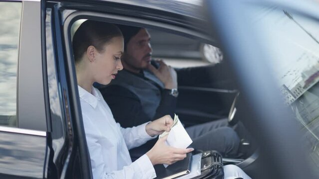 Side view portrait of slim young beautiful woman counting cash sitting in car with blurred man. Caucasian concentrated industrial spy with money bribe from businessman in vehicle downtown