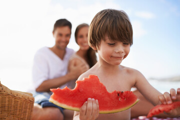 Juicy and sweet. A happy little boy biting into a watermelon with his parents looking on.