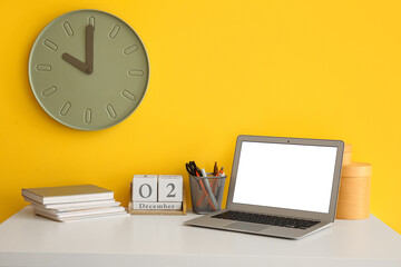 Modern laptop, cube calendar and stationery supplies on table near color wall with big clock