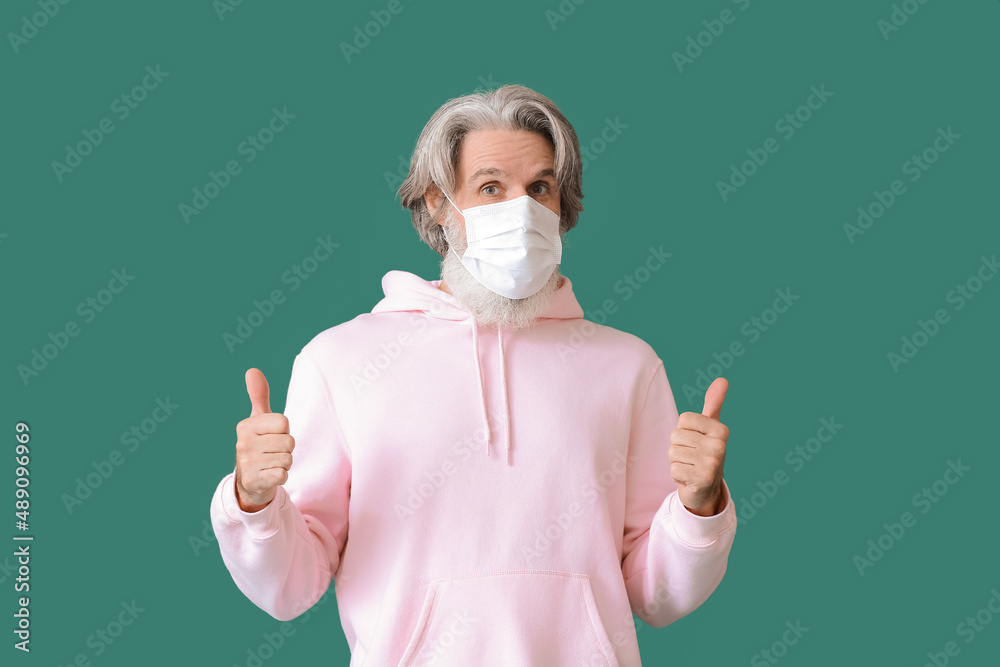 Poster senior man in medical mask showing thumbs-up on green background