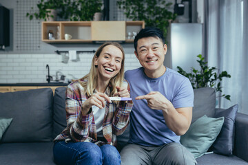 Young happy asian family and woman looking at camera and rejoicing smiling, getting a positive pregnancy test, sitting at home on the couch