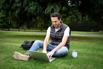 Caucasian man sitting on grass working with laptop at a park. Middle aged man working outdoors.