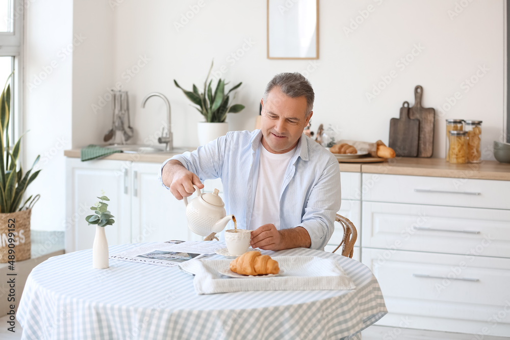 Sticker senior man pouring tea into cup at table in kitchen