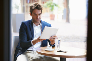 Catching up on his coffee break. Shot of a young businessman enjoying a cup of coffee while using a digital tablet.