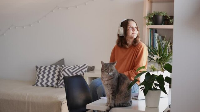 Teenage girl sitting on top of table in her room relaxing looking out the window, resting after studying at home online class talking to teacher via video call 
