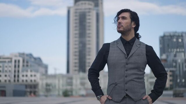Medium shot portrait of elegant confident businessman in waistcoat putting hands in pockets looking around in slow motion. Successful Caucasian man standing on urban city square outdoors