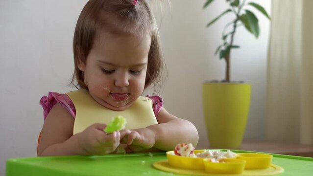 Close up of lovely baby girl eating have breakfast eggs vegetables sausage by herself at home. Adorable possitive happy toddler child eats with dirty face at table indoor. Food, nutrition healthy life