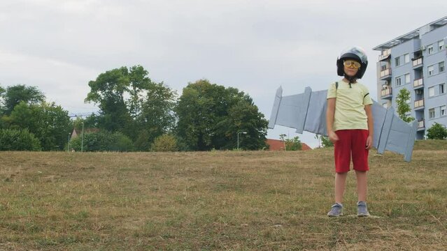 Kid boy dressed as pilot plays with handmade paper wings outdoor