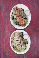 Caesar salads. Top view of two Caesar salads with lettuce, parmesan cheese, bread croutons, bacon and Caesar dressing. One with grilled chicken breast and the other one with smoked salmon.