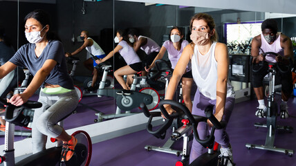 Men and women wearing protective masks ride stationary bike in a fitness club