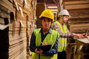 Female worker in safety uniform and hard hat, supervisor quality inspects packaging stock order supply at factory storage warehouse, piles of stacking paper manufacture, recycling production industry.