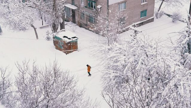 An Old Rusty Garage Stands in a Snow-Covered Yard of a Residential Brick House. Top view. A person passing by. The yard is surrounded by snow-covered trees. Winter windy weather. Winter landscape.