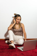 Adult blonde woman sitting on a red carpet, having fun while using her headphones and her laptop listening music