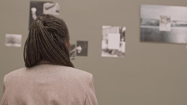 Rear-view waist-up slowmo shot of young African-American woman looking at black-and-white photos on wall at exhibition in modern art gallery