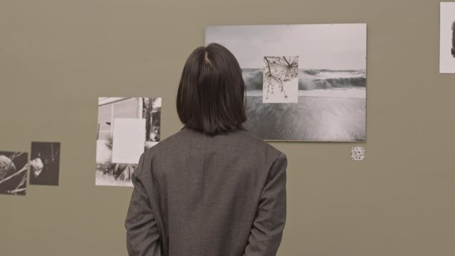 Rear-view medium slowmo shot of long-haired man examining abstract black and white photos on wall of contemporary art gallery