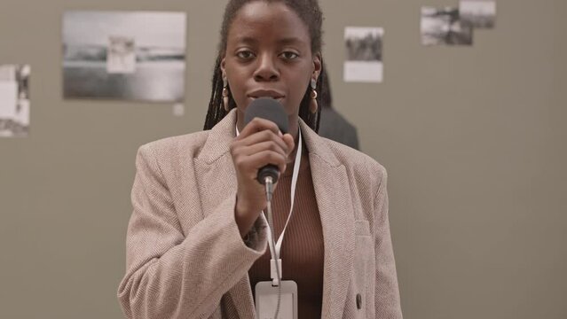 Waist-up slowmo shot of young African-American female guide looking at camera while speaking in microphone about abstract photos in contemporary art gallery