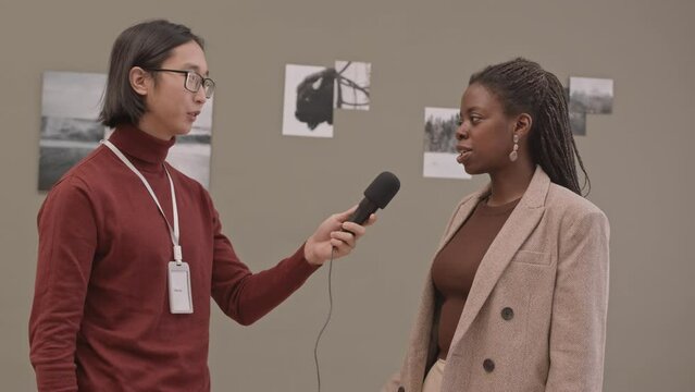 Medium slowmo shot of young Asian male gallerist with microphone taking interview of beginning African-American female photographer whose works hanging on wall of modern art gallery