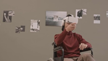 Medium slowmo shot of young Asian man with disability in wheelchair wearing VR headset during virtual art exhibition in museum