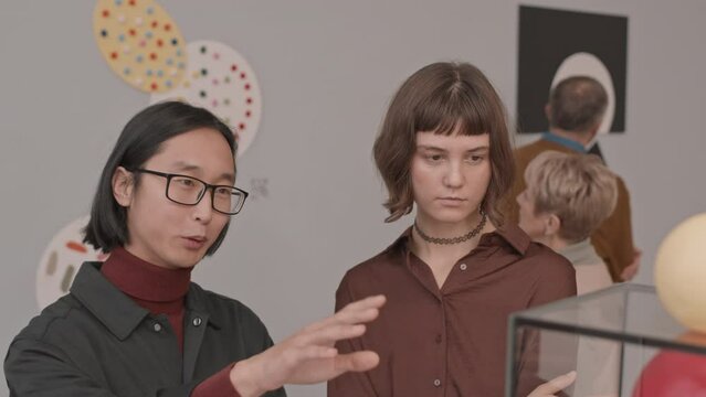 Medium slowmo shot of young Asian man and Caucasian woman discussing conceptual art object while visiting exhibition in museum of modern arts
