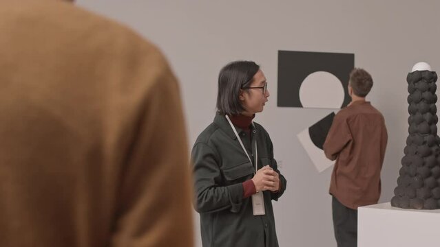 Medium slowmo shot of young Asian male guide talking to tourists visiting exhibition in museum of contemporary arts