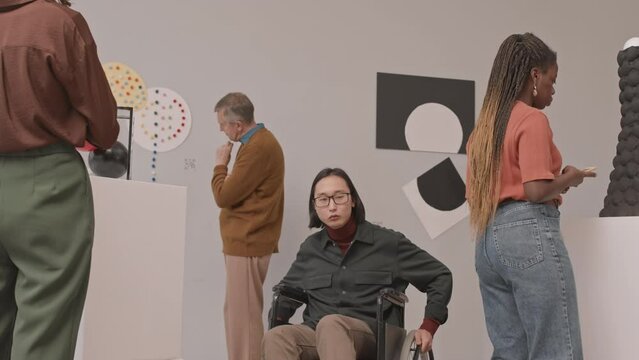 Medium slowmo portrait of young Asian man with disability in wheelchair visiting exhibition at museum of contemporary arts then approaching to camera