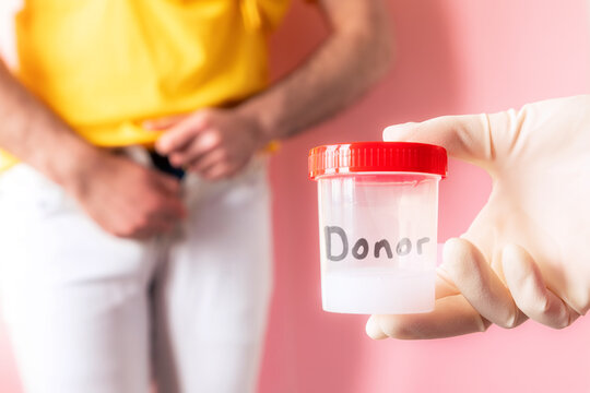 Doctor's hand in a medical glove holds a plastic jar with a sample of sperm. In the defocused background a man shyly zips up his jeans. The concept of sperm donation and artificial insemination