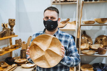 A white man wearing a mask shows off his wares at a craft show. A man holds a round wooden plate in his hands. Carpenter is handmade.