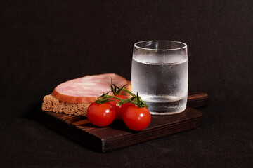 A glass of vodka, a sandwich with rye bread and ham and red cherry tomatoes