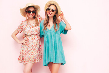 Two young beautiful smiling brunette hipster female in trendy summer dresses. Sexy carefree women posing near pink wall. Positive models having fun. Cheerful and happy. In hats and sunglasses