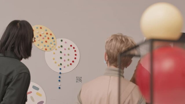 Rear-view slowmo shot of mature Caucasian couple listening to Asian male guide while looking at conceptual art object hanging on wall in museum of modern arts