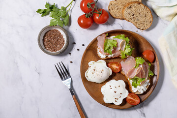 Easter breakfast or brunch. Delicious breakfast or snack - poached egg and cream cheese toast whole grain rye bread, prosciutto, arugula on a marble tabletop. Top view flat lay. Copy space.