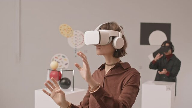 Medium slowmo shot of young woman in vr headset moving her hands during virtual reality exhibition in contemporary art museum