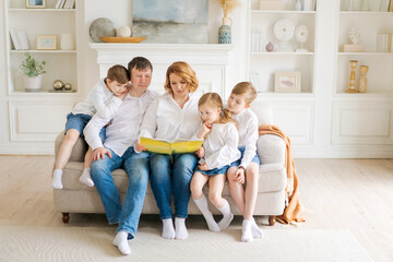 Happy family reading book at home. Mom and dad and young children sit together on a cozy sofa in a new large, bright living room with white walls, furniture and shelves in the background.