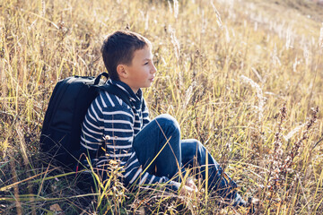 Naklejka na ściany i meble Autumn portrait of teenager boy sitting on grass. Young man with backpack resting relaxing, contemplating, thinking, daydreaming. Teen deep in thought Local travel. Active lifestyle People from behind