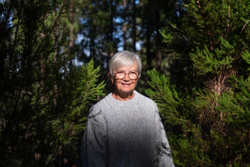 Naklejka na ściany i meble Portrait of attractive smiling caucasian senior woman in the woods. Smiling mature gray haired elderly people enjoying nature, care of environment, freedom vacation travel in the mountain forest