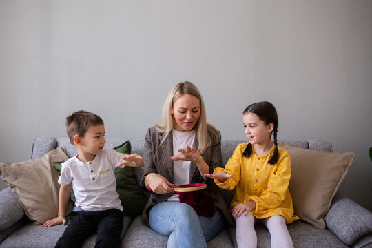 Happy Mom And Kids Are Sitting On A Gray Sofa In The Room And Playing Tricks With The Kids