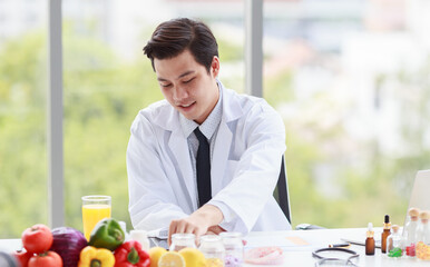 Portrait studio shot Asian male professional nutritionist dietitian doctor in white lab coat and stethoscope sitting holding pen smiling at working desk full of fruits vegetable nutrition supplements
