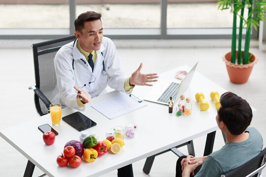 Top view shot Asian male professional nutritionist dietitian doctor in white lab coat and stethoscope sitting smiling discussing with patient at working desk about nutrition food and supplement pill