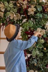 A child in a straw hat and a blue shirt in front of a flower arrangement. View from the back. The boy touches a white flower. Spring concept. Happy childhood.