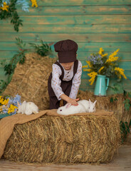 A child strokes white rabbits against a floral blue wall. A boy in a brown cap and overalls with fluffy animals sits on the hay. Easter concept. Happy childhood.