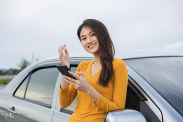 A young Asian woman drives a beautiful nature drive. she was standing in front of the car on the roadside. she uses the smartphone to call services.