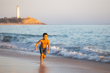 Little kid running on the beach with a lighthouse background - obrazy, fototapety, plakaty